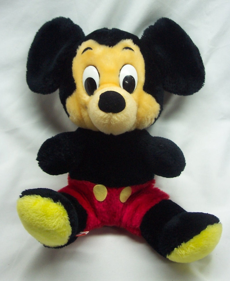 May include: A plush black and yellow Mickey Mouse toy wearing red shorts with yellow buttons. The toy has large black ears and a black nose.