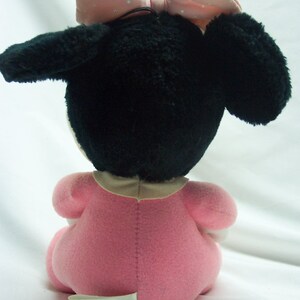 May include: A plush toy of a baby Minnie Mouse wearing a pink dress and a pink bow with white polka dots. The toy is sitting on a white surface. The toy has a label on the bottom that says "Made in Korea."