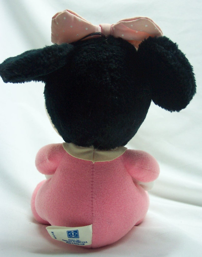May include: A plush toy of a baby Minnie Mouse wearing a pink dress and a pink bow with white polka dots. The toy is sitting on a white surface. The toy has a label on the bottom that says "Made in Korea."