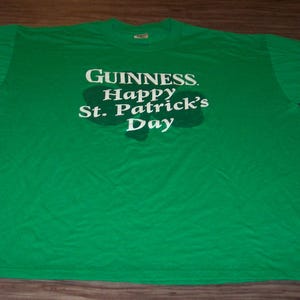 May include: A green t-shirt with the Guinness logo and the text "Happy St. Patrick's Day" in white. The text is centered on the front of the shirt, above a shamrock shape. The shirt is laid flat on a wooden surface.