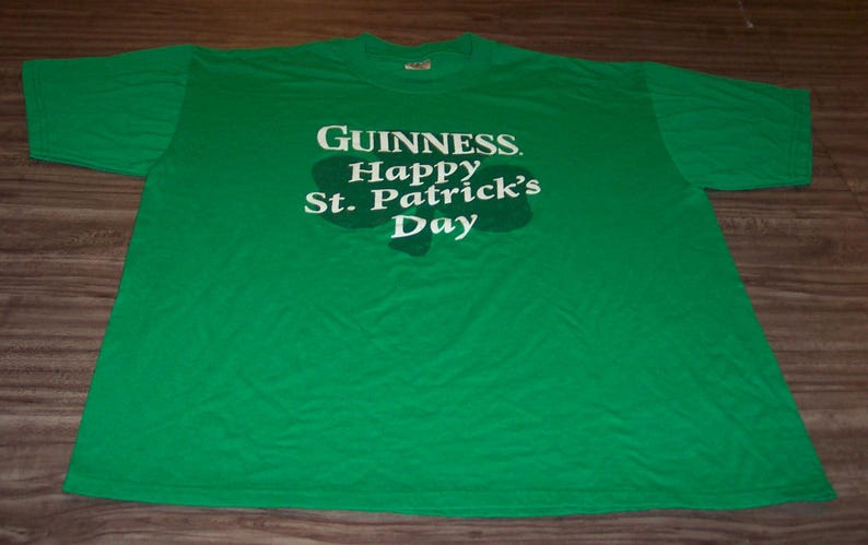 May include: A green t-shirt with the Guinness logo and the text "Happy St. Patrick's Day" in white. The text is centered on the front of the shirt, above a shamrock shape. The shirt is laid flat on a wooden surface.