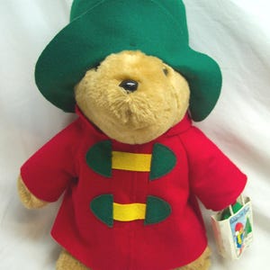 May include: A plush Paddington Bear toy wearing a green hat and a red coat with green and yellow accents. The bear is holding a small tag with an illustration.