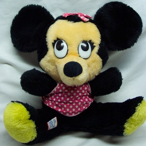 May include: A plush black and yellow Minnie Mouse toy wearing a pink and white polka dot swimsuit. The toy has large black eyes and a pink bow.