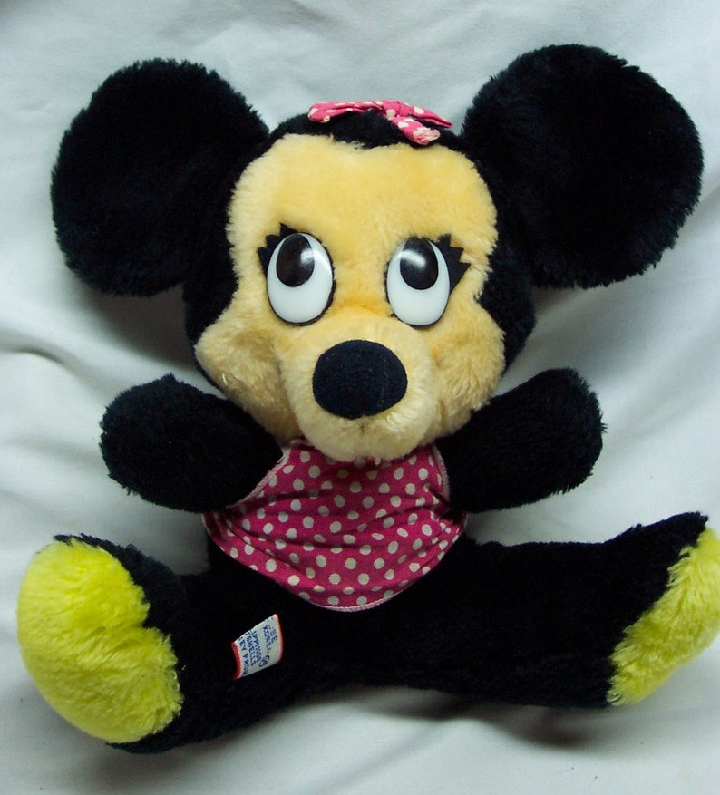 May include: A plush black and yellow Minnie Mouse toy wearing a pink and white polka dot swimsuit. The toy has large black eyes and a pink bow.