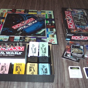 以下が含まれることがあります： スター・ウォーズのキャラクターと宇宙船をフィーチャーしたモノポリーボードゲーム。ゲームボードが開いており、ゲームピース、お金、チャンスとコミュニティチェストカードが表示されています。ゲームの箱も表示されています。