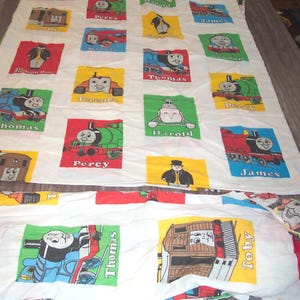 May include: White bed sheet featuring colorful squares with illustrations of Thomas the Tank Engine and friends. Each square has a different character, including Thomas, Percy, James, and Harold, with their names printed below. The sheet is likely for a child's bed.