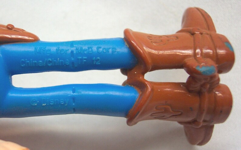 Pu&ograve; includere: Primo piano delle gambe di un giocattolo da cowboy in plastica blu con stivali marroni. L'iscrizione sulle gambe recita "Mfg for Mel Corp. China/China TF 12" e "&copy; Disney".