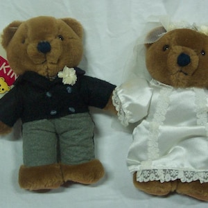 May include: A pair of brown teddy bears dressed in wedding attire. The groom bear is wearing a black suit with a white flower on his lapel. The bride bear is wearing a white satin dress with a lace trim and a veil. The bears are holding a bouquet of flowers.