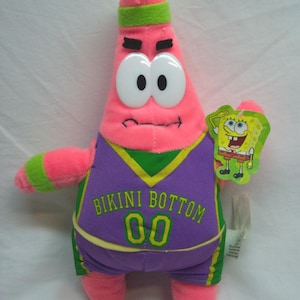 May include: Pink plush Patrick Star doll wearing a purple and green basketball jersey with "BIKINI BOTTOM 00" printed on it. The doll has large white eyes, a green headband, and a SpongeBob SquarePants tag.