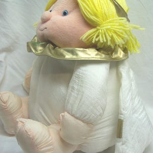 May include: A plush angel doll with yellow yarn hair and a gold halo. The doll has a light pink face, arms, and feet, and is wearing a white dress with gold trim. The doll is sitting down.