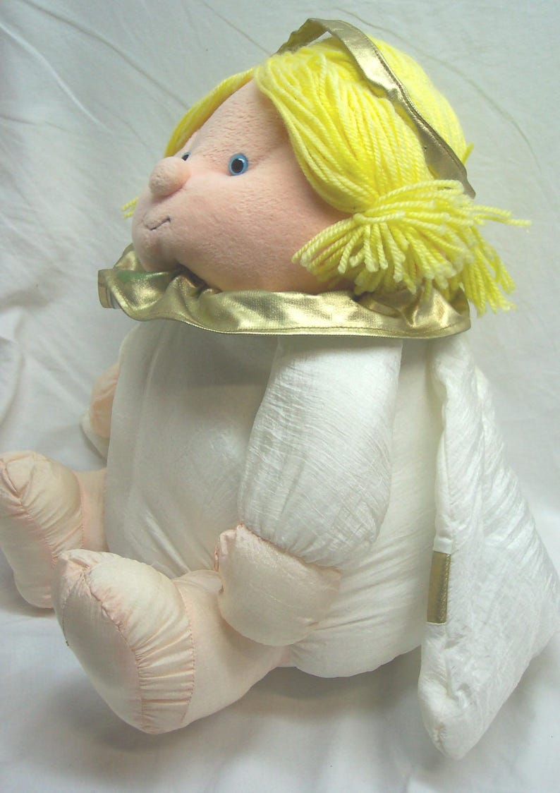 May include: A plush angel doll with yellow yarn hair and a gold halo. The doll has a light pink face, arms, and feet, and is wearing a white dress with gold trim. The doll is sitting down.
