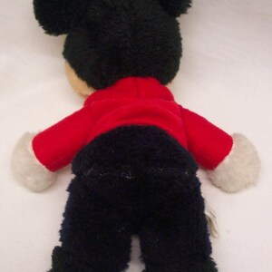 May include: A plush toy of a cartoon character, viewed from the back. The toy has large black ears, a red jacket, black pants, and white gloves. The toy is on a white background.