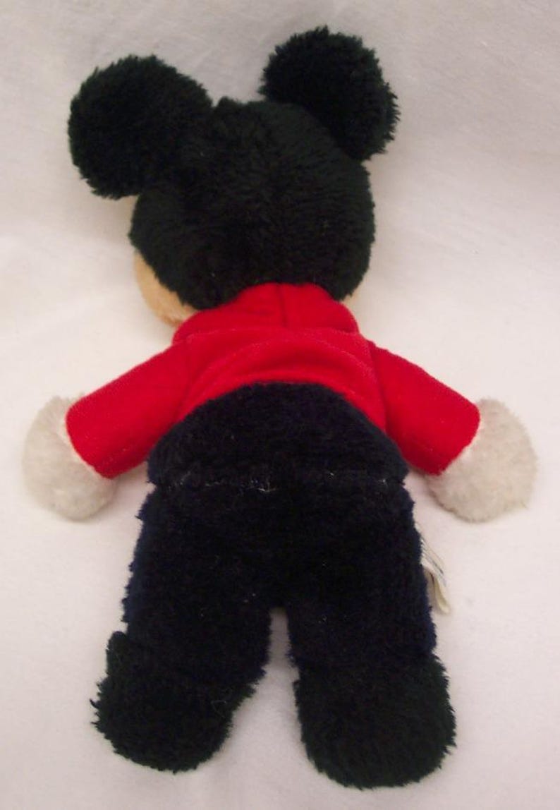 May include: A plush toy of a cartoon character, viewed from the back. The toy has large black ears, a red jacket, black pants, and white gloves. The toy is on a white background.