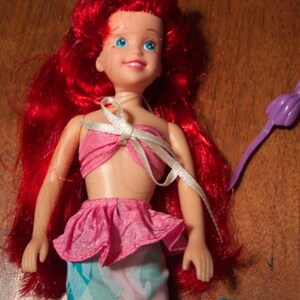 May include: A red-haired doll wearing a pink and blue mermaid costume. The doll has blue eyes and a white ribbon tied around her neck.
