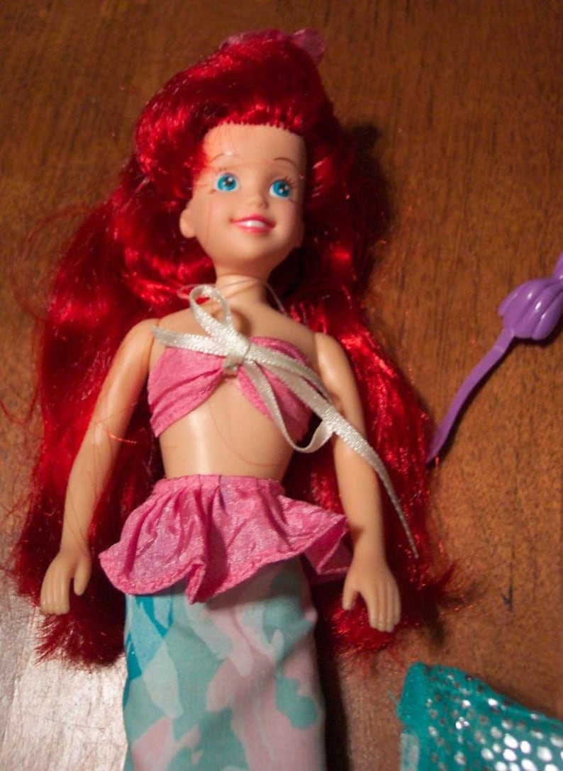 May include: A red-haired doll wearing a pink and blue mermaid costume. The doll has blue eyes and a white ribbon tied around her neck.