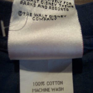May include: A white fabric clothing tag with red trim on a dark blue garment. The text reads: MADE EXCLUSIVELY FOR THE WALT DISNEY THEME PARKS AND RESORTS, THE WALT DISNEY COMPANY, 100% COTTON, MACHINE WASH SEPARATE, LUKEWARM WATER, NON-CHLORINE BLEACH.