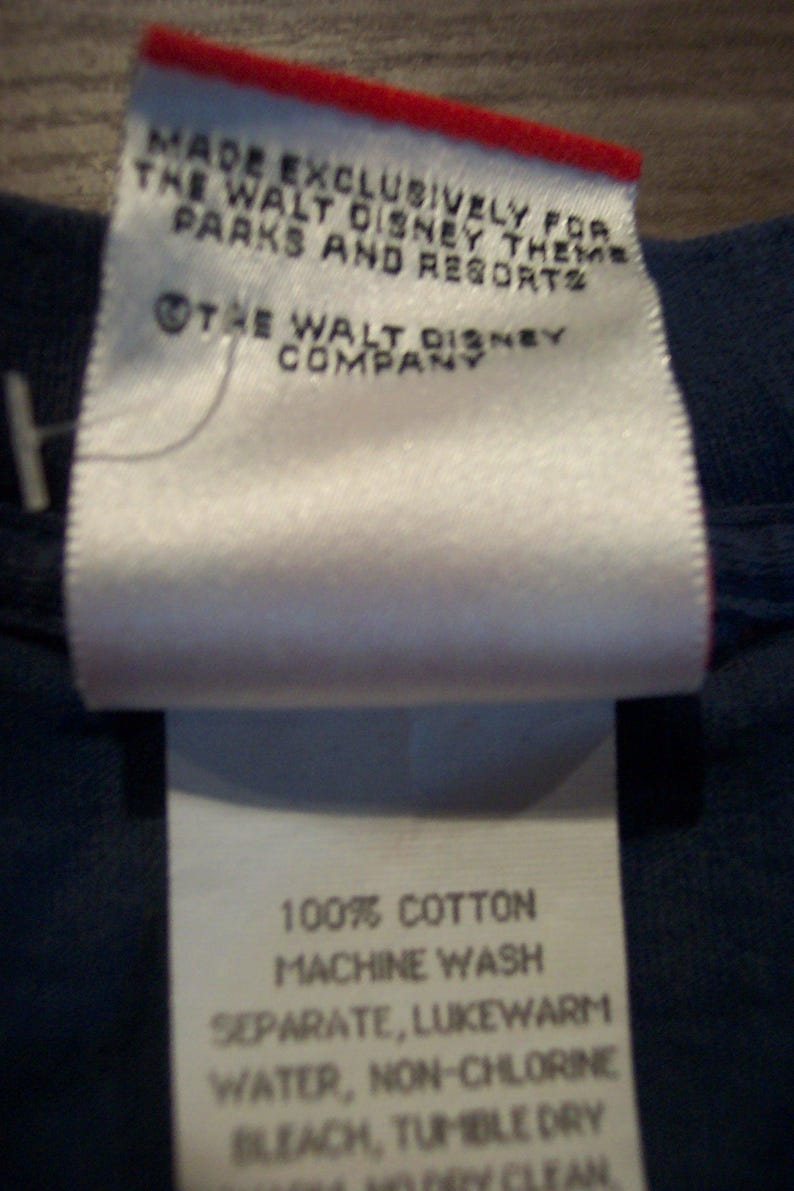 May include: A white fabric clothing tag with red trim on a dark blue garment. The text reads: MADE EXCLUSIVELY FOR THE WALT DISNEY THEME PARKS AND RESORTS, THE WALT DISNEY COMPANY, 100% COTTON, MACHINE WASH SEPARATE, LUKEWARM WATER, NON-CHLORINE BLEACH.
