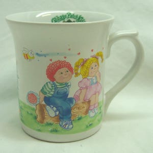 May include: White ceramic mug featuring Cabbage Patch Kids characters. The mug has a curved handle and is decorated with illustrations of a boy and girl sitting on a log, with a bee and hearts. The mug has the words "Cabbage Patch Kids" printed on the rim.