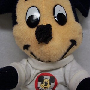 K&ouml;nnte beinhalten: Ein Pl&uuml;sch-Mickey-Mouse-Spielzeug, das ein wei&szlig;es Hemd mit einem roten Kreis und dem Text "Mickey Mouse Club" tr&auml;gt. Das Spielzeug hat schwarze Ohren, eine schwarze Nase und gro&szlig;e schwarze Augen.