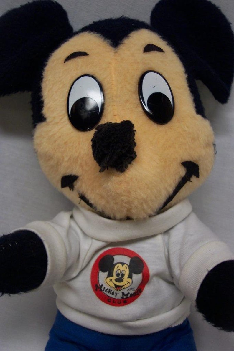 K&ouml;nnte beinhalten: Ein Pl&uuml;sch-Mickey-Mouse-Spielzeug, das ein wei&szlig;es Hemd mit einem roten Kreis und dem Text "Mickey Mouse Club" tr&auml;gt. Das Spielzeug hat schwarze Ohren, eine schwarze Nase und gro&szlig;e schwarze Augen.