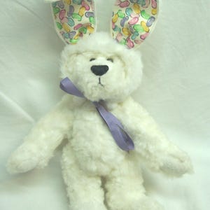 May include: A white plush bunny with a lavender ribbon around its neck. The bunny's ears are lined with a colorful jelly bean pattern. The bunny has a black nose and a soft, fluffy texture.