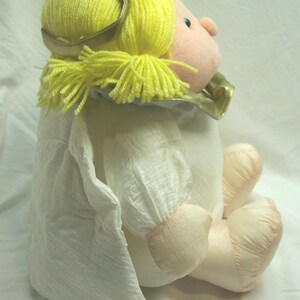 May include: A plush angel doll with yellow yarn hair and a gold halo. The doll has a pink face, arms, and legs, and is wearing a white dress with a ruffled collar and sleeves. The doll is sitting down.