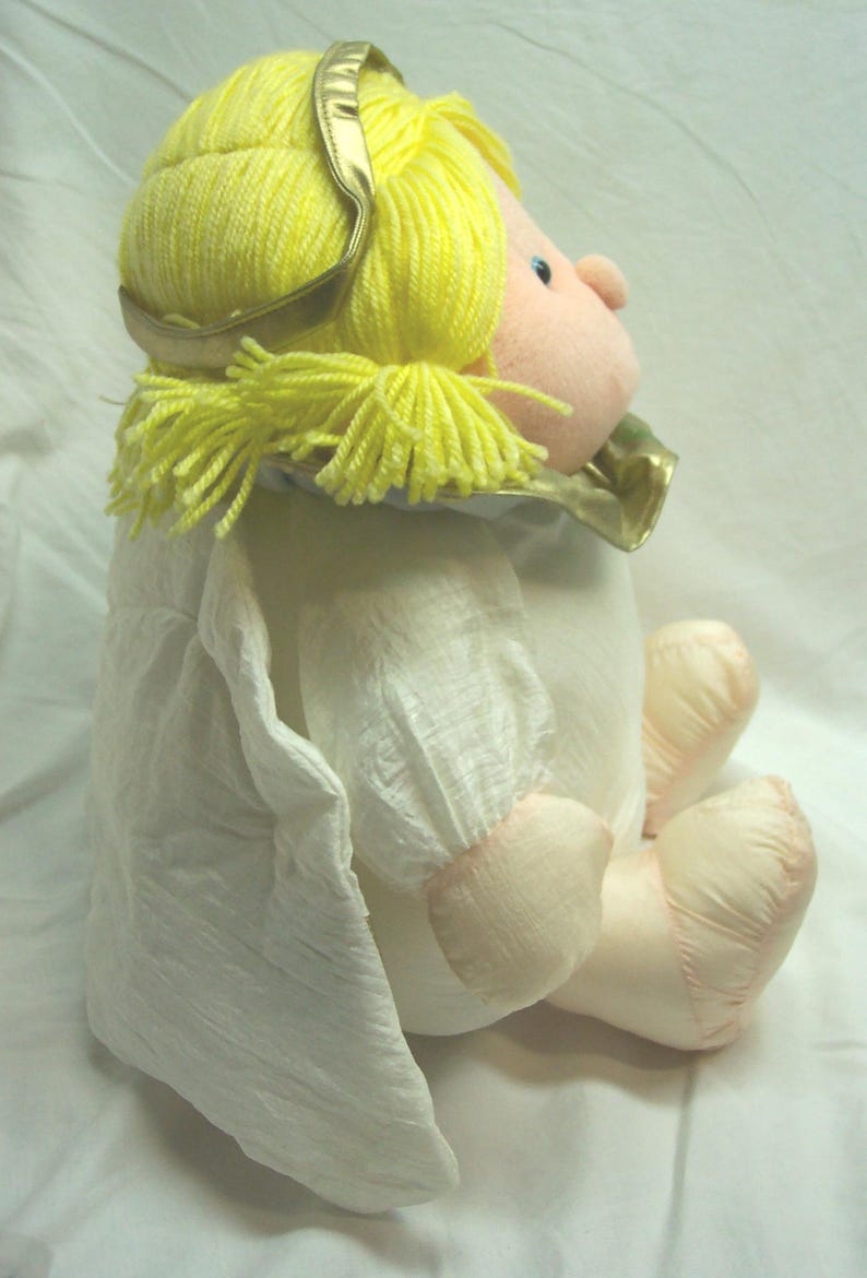 May include: A plush angel doll with yellow yarn hair and a gold halo. The doll has a pink face, arms, and legs, and is wearing a white dress with a ruffled collar and sleeves. The doll is sitting down.