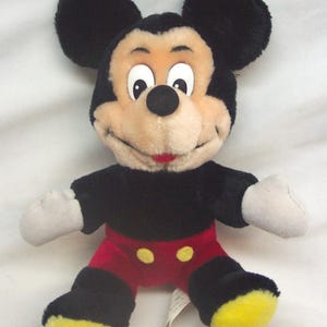 Pode incluir: Um brinquedo de pel&uacute;cia do Mickey Mouse com orelhas pretas, corpo preto, cal&ccedil;&otilde;es vermelhos com bot&otilde;es amarelos e p&eacute;s amarelos. O brinquedo tem um rosto bege, luvas brancas e uma express&atilde;o sorridente.