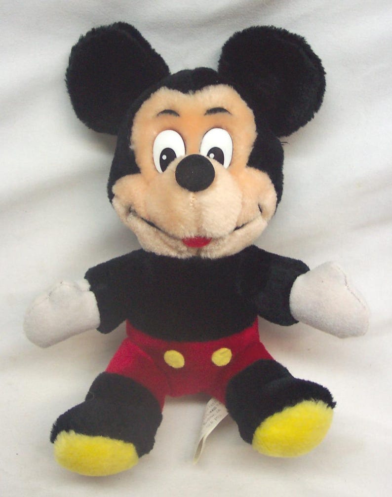 Pode incluir: Um brinquedo de pel&uacute;cia do Mickey Mouse com orelhas pretas, corpo preto, cal&ccedil;&otilde;es vermelhos com bot&otilde;es amarelos e p&eacute;s amarelos. O brinquedo tem um rosto bege, luvas brancas e uma express&atilde;o sorridente.