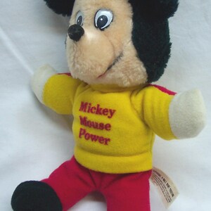 May include: A plush Mickey Mouse doll with black ears, a tan face, and a yellow shirt with the text "Mickey Mouse Power." The doll wears red pants and black shoes. A classic Disney collectible.