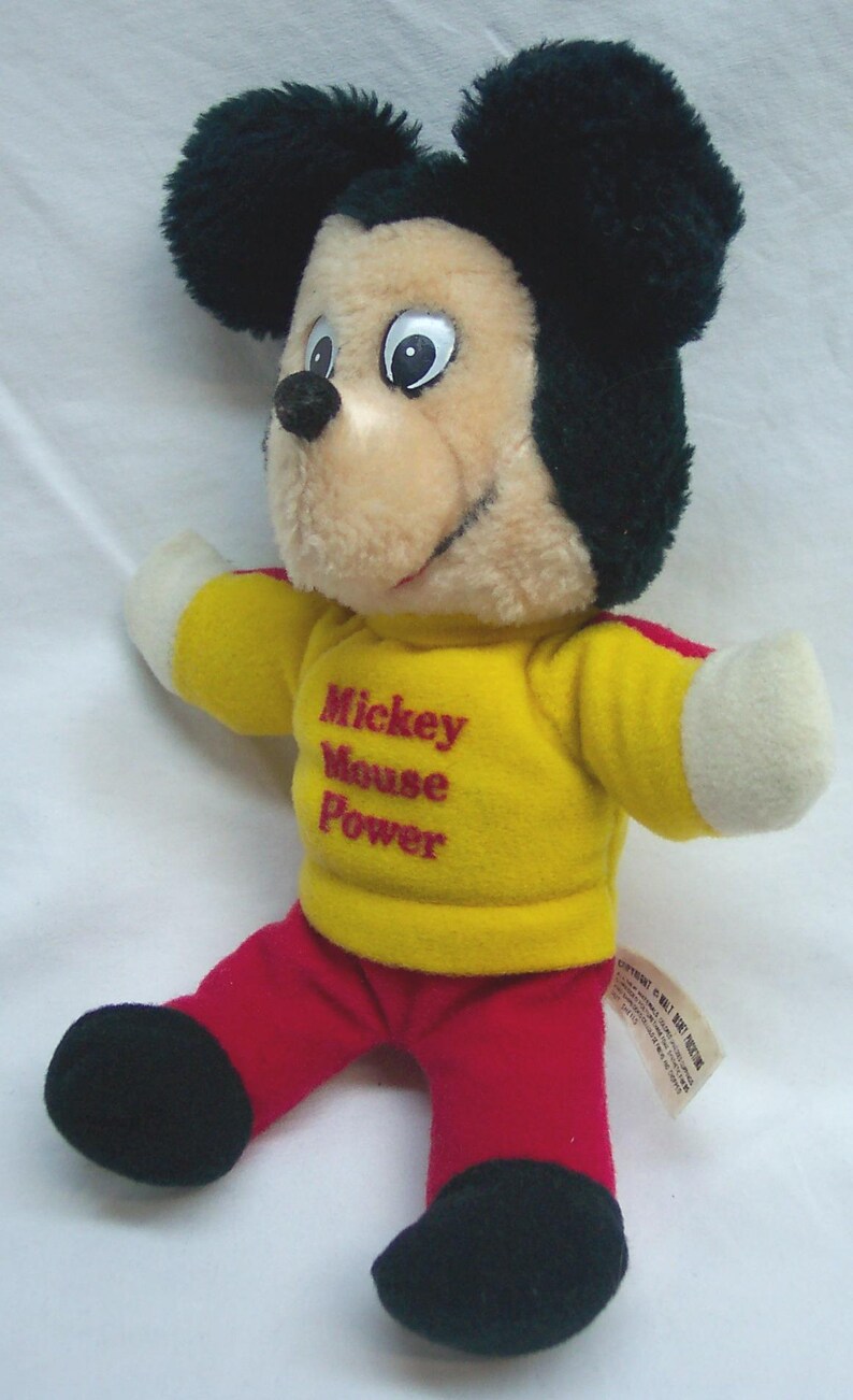 May include: A plush Mickey Mouse doll with black ears, a tan face, and a yellow shirt with the text "Mickey Mouse Power." The doll wears red pants and black shoes. A classic Disney collectible.
