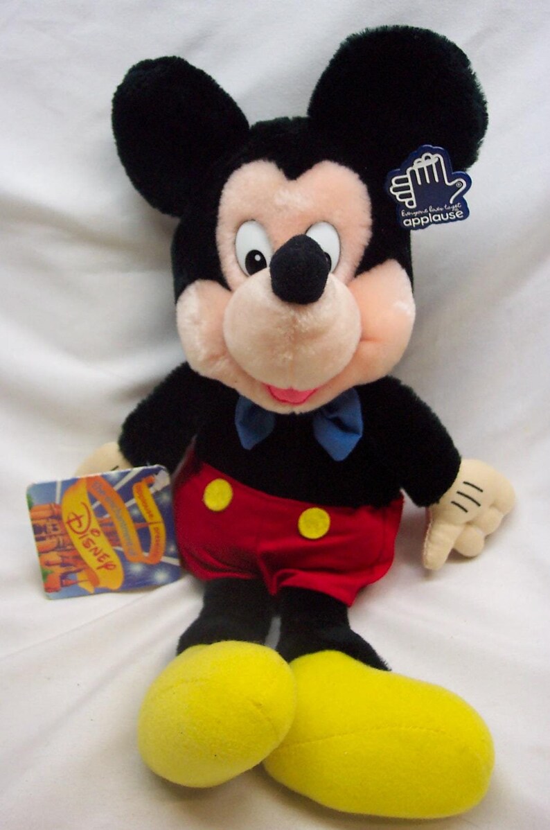 May include: A plush Mickey Mouse toy wearing red shorts with yellow buttons, a black shirt with a blue bow tie, and yellow shoes. The toy has a black nose and large, round ears. The toy is holding a tag that says "Applause".
