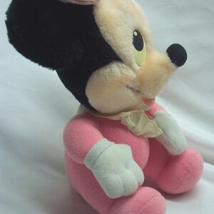 May include: A pink plush toy of Minnie Mouse wearing a pink bow and a pink dress. The toy is sitting on a white surface.