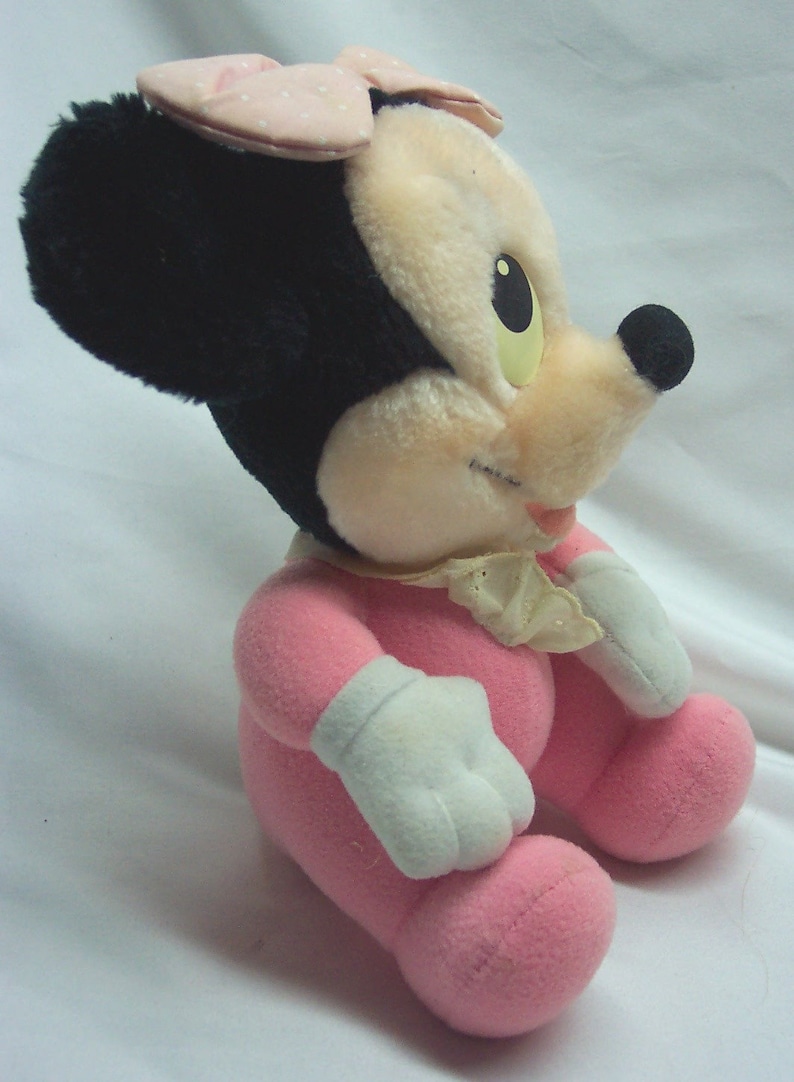 May include: A pink plush toy of Minnie Mouse wearing a pink bow and a pink dress. The toy is sitting on a white surface.
