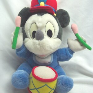 Puede incluir: Un juguete de peluche de Mickey Mouse con un sombrero rojo y azul y un mono azul, sosteniendo baquetas y sentado en un tambor amarillo y rojo.