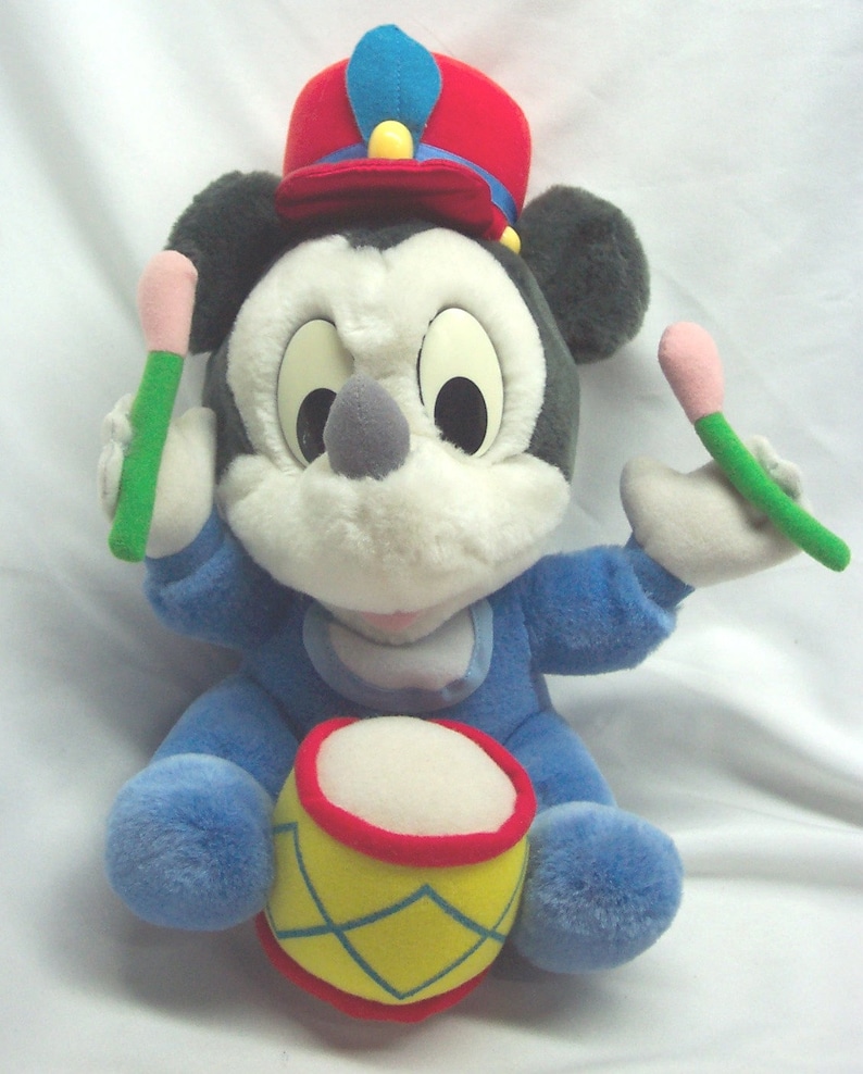 Puede incluir: Un juguete de peluche de Mickey Mouse con un sombrero rojo y azul y un mono azul, sosteniendo baquetas y sentado en un tambor amarillo y rojo.