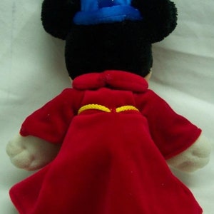 May include: A plush Mickey Mouse dressed as a sorcerer. He is wearing a blue hat with white stars and a red robe with a yellow sash.