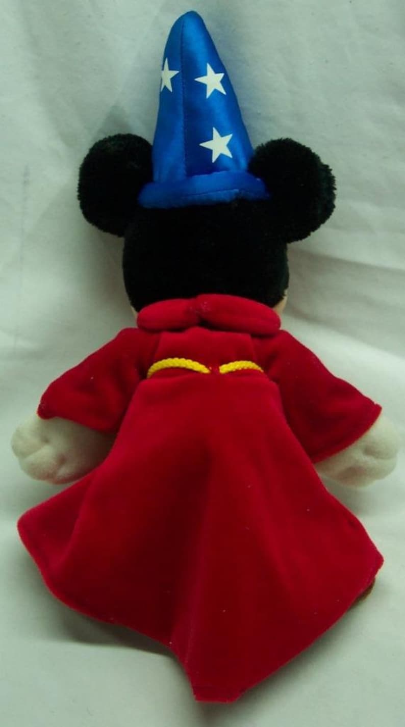 May include: A plush Mickey Mouse dressed as a sorcerer. He is wearing a blue hat with white stars and a red robe with a yellow sash.