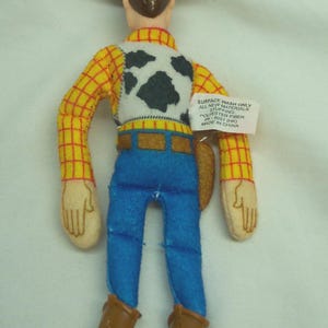 Puede incluir: Un juguete de peluche de un personaje de vaquero, con sombrero marr&oacute;n, camisa a cuadros amarillos, vaqueros azules y botas marrones. El juguete tiene un chaleco blanco con manchas negras y manos color beige.