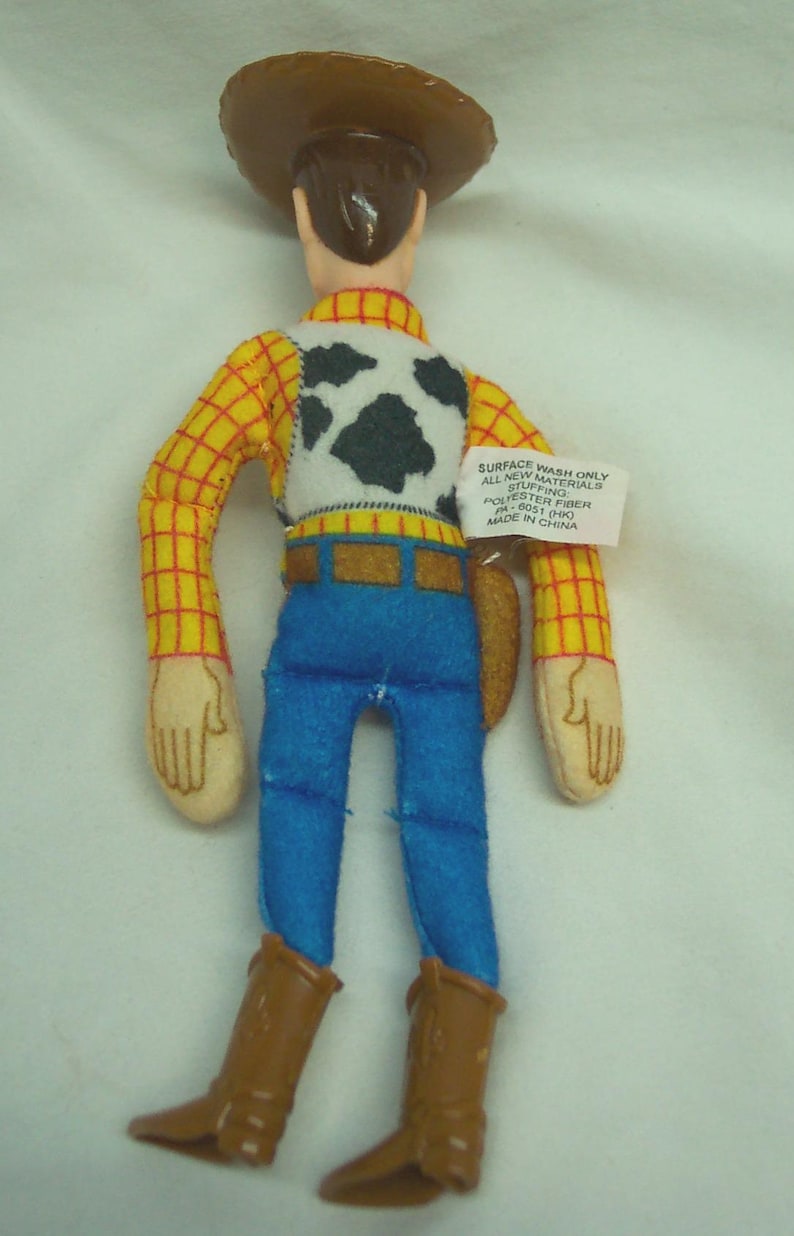 Puede incluir: Un juguete de peluche de un personaje de vaquero, con sombrero marr&oacute;n, camisa a cuadros amarillos, vaqueros azules y botas marrones. El juguete tiene un chaleco blanco con manchas negras y manos color beige.