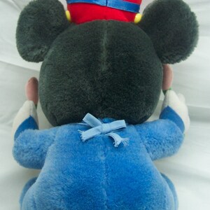 May include: A plush Mickey Mouse toy wearing a blue and white outfit with a red hat and a blue bow. The toy is sitting on a white surface.