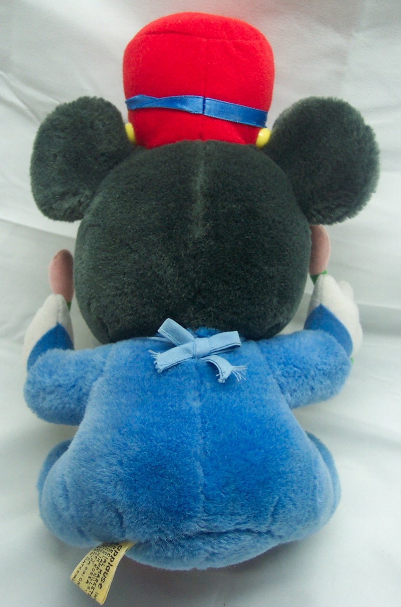 May include: A plush Mickey Mouse toy wearing a blue and white outfit with a red hat and a blue bow. The toy is sitting on a white surface.
