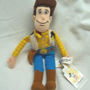 May include: A plush toy of Woody from Toy Story, wearing a brown cowboy hat, yellow plaid shirt, blue jeans, and brown boots. The toy has a white vest and a sheriff's badge. A Disney World tag is attached.