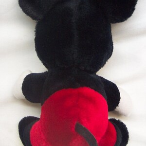 Pode incluir: Um brinquedo de pel&uacute;cia do Mickey Mouse, visto por tr&aacute;s. O brinquedo apresenta grandes orelhas pretas, um corpo preto, luvas brancas e cal&ccedil;&otilde;es vermelhos. A cauda &eacute; preta. O brinquedo est&aacute; sobre uma superf&iacute;cie branca.