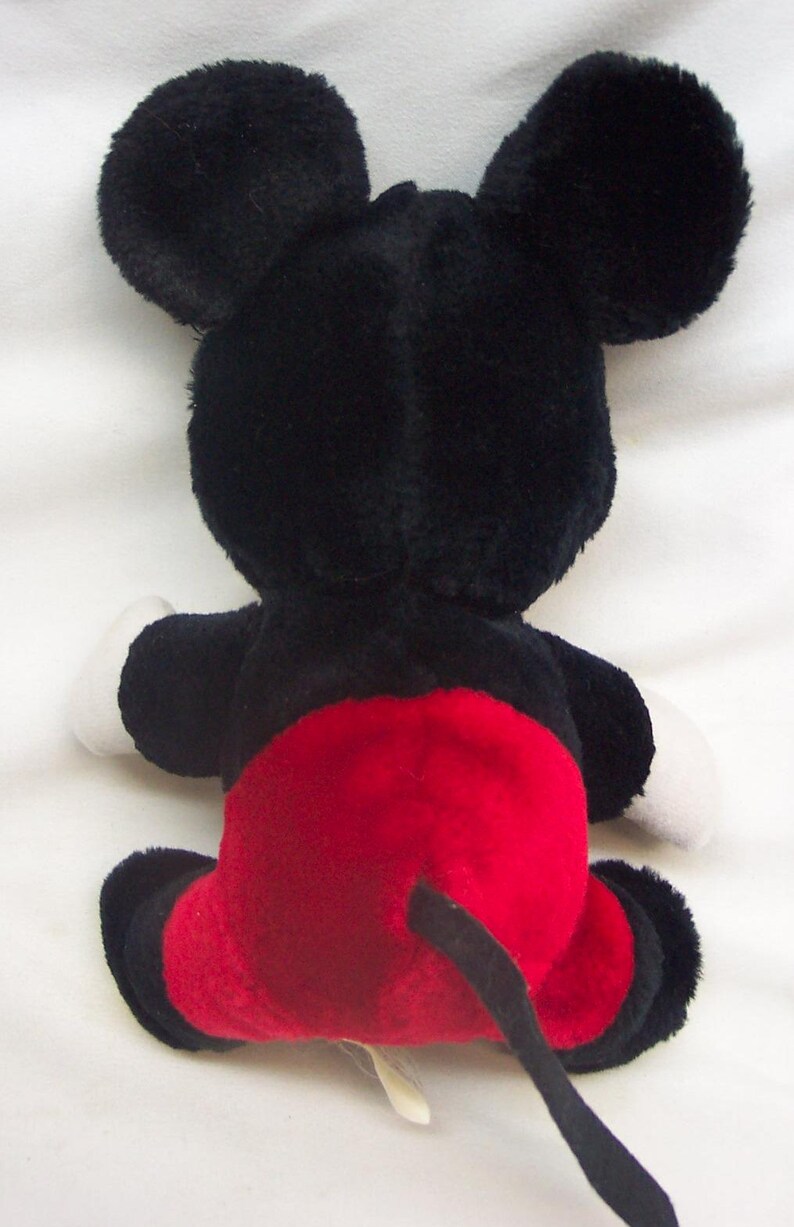 Pode incluir: Um brinquedo de pel&uacute;cia do Mickey Mouse, visto por tr&aacute;s. O brinquedo apresenta grandes orelhas pretas, um corpo preto, luvas brancas e cal&ccedil;&otilde;es vermelhos. A cauda &eacute; preta. O brinquedo est&aacute; sobre uma superf&iacute;cie branca.