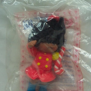 May include: A small plastic doll with black hair, wearing a red dress with yellow buttons and a white and red candy cane. The doll is wearing blue shoes and is packaged in a clear plastic bag with a pink and white background.