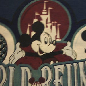 May include: A dark blue t-shirt featuring a graphic of Mickey Mouse with arms outstretched in front of a castle silhouette. The text reads WORLD REUNION and WALT DISNEY WORLD 25TH ANNIVERSARY, with a small globe icon replacing the letter O.