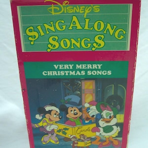 May include: A vintage Disney's Sing Along Songs VHS box. The box is pink and green with the title "Disney's Sing Along Songs" in large yellow letters. The bottom of the box features a cartoon of Mickey, Minnie, Donald, and Daisy Duck singing Christmas carols.