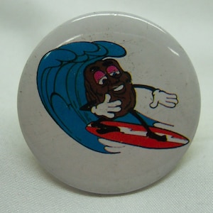 May include: A white button with a cartoon brown raisin surfing on a blue wave. The raisin is wearing a white glove and has a pink tongue sticking out. The wave is breaking and the raisin is riding a red surfboard.