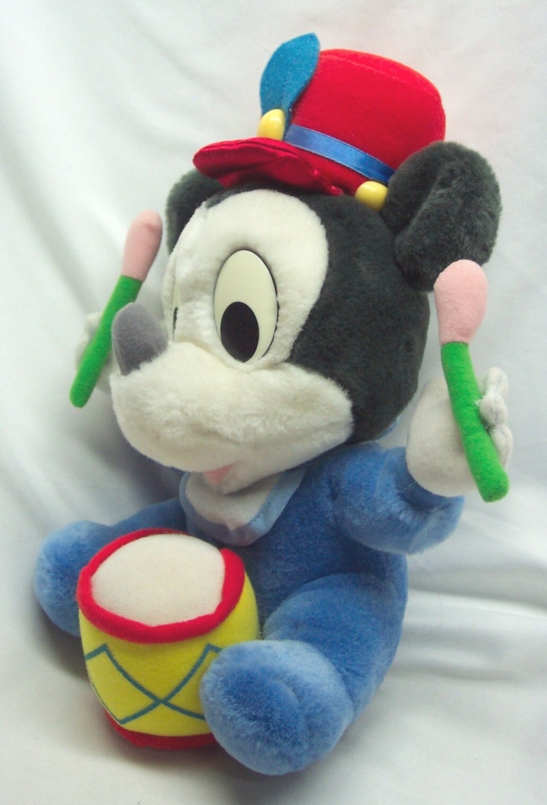 Puede incluir: Un juguete de peluche de Mickey Mouse con un sombrero rojo y azul, un mono azul y sosteniendo dos baquetas. El juguete est&aacute; sentado en un tambor amarillo y rojo.